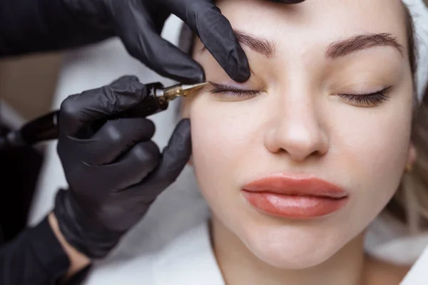 Primer plano de una mujer, en un salón de belleza con los ojos cerrados, un cosmetólogo hace maquillaje permanente delineador de ojos. Profesional en guantes negros y con un especial también — Foto de Stock