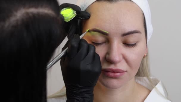 De hand van een make-up artiest in zwarte handschoenen brengt een gele wenkbrauwpasta aan op de wenkbrauw. Professionele stijlvolle permanente make-up, wenkbrauwkleuring. Sluiten.. — Stockvideo