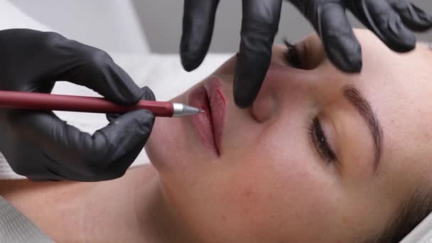 Jonge vrouw met permanente make-up op haar lippen in een kosmetologen salon. Permanente make-up tatoeage. trek een lijn met een rood lippotlood — Stockvideo