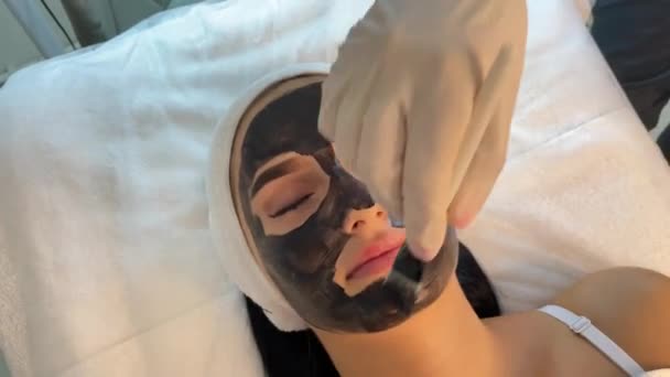 Cosmetólogo aplicando máscara negra en cara de mujer bonita usando guantes negros, mujer hermosa en spa teniendo procedimientos faciales — Vídeo de stock