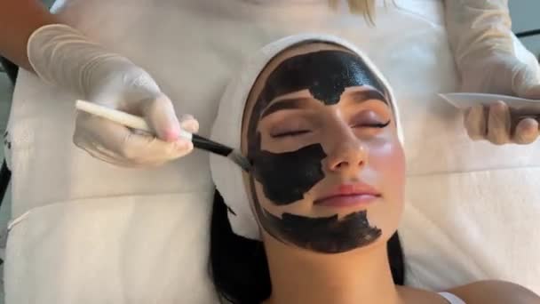 Κοσμητολόγος εφαρμογή μαύρη μάσκα σε όμορφη γυναίκα πρόσωπο φορώντας μαύρα γάντια, πανέμορφη γυναίκα σε spa έχοντας διαδικασίες του προσώπου — Αρχείο Βίντεο