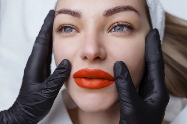 Stały makijaż. Tatuaże na ustach. Jasnoczerwone seksowne usta w rękach kosmetologa w czarnych rękawiczkach — Zdjęcie stockowe