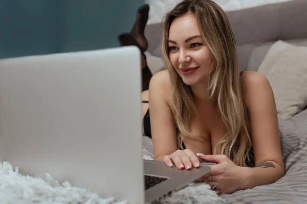 黒いランジェリーのセクシーな女性はベッドの上にあり、ラップトップを使用しています。ウェブカメラ上のセックスモデルの概念と出会い系サイト — ストック写真