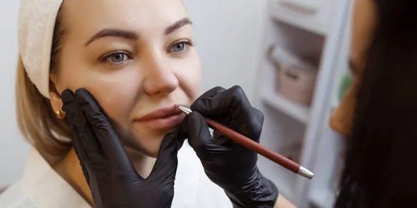 Mujer joven con maquillaje permanente en los labios en un salón de cosmetólogos. Tatuaje maquillaje permanente. dibujar una línea con un lápiz labial rojo — Foto de Stock