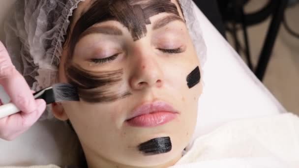 美容師は美しい女性の顔に黒いマスクを適用します。スパサロンで顔のための炭素剥離手順を行う豪華な女性. — ストック動画