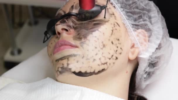 美容師は美しい女性の顔に黒いマスクを適用します。スパサロンで顔のための炭素剥離手順を行う豪華な女性. — ストック動画