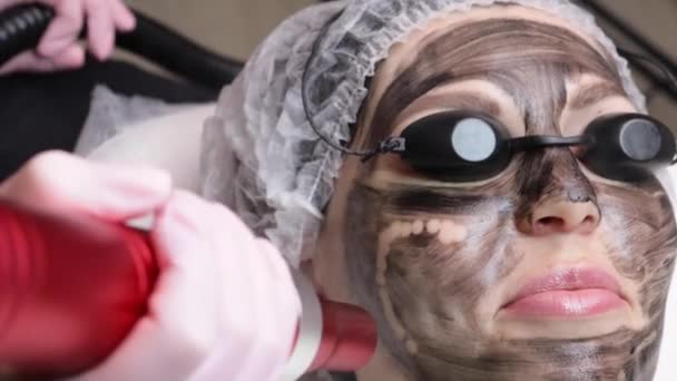 Kosmetolog applicerar svart mask på ansiktet av vacker kvinna. vacker kvinna i spa salong gör kol peeling förfaranden för ansiktet. — Stockvideo