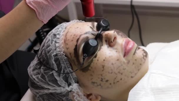 Beautician applies black mask on face of beautiful woman. gorgeous woman in spa salon doing carbon peeling procedures for face. — Stock Video