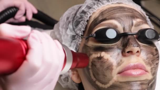 Αισθητικός εφαρμόζει μαύρη μάσκα στο πρόσωπο της όμορφης γυναίκας. πανέμορφη γυναίκα στο σπα σαλόνι κάνει διαδικασίες ξεφλούδισμα άνθρακα για το πρόσωπο. — Αρχείο Βίντεο