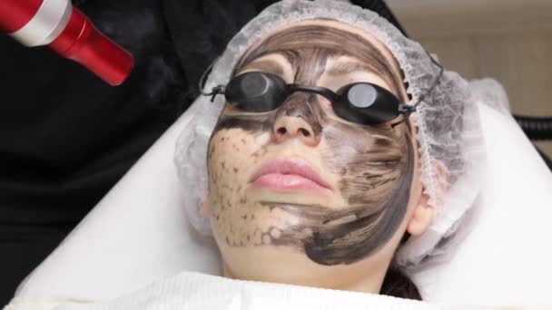 Αισθητικός εφαρμόζει μαύρη μάσκα στο πρόσωπο της όμορφης γυναίκας. πανέμορφη γυναίκα στο σπα σαλόνι κάνει διαδικασίες ξεφλούδισμα άνθρακα για το πρόσωπο. — Αρχείο Βίντεο