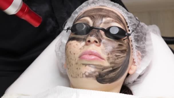 Esteticista aplica máscara negra en la cara de la mujer hermosa. hermosa mujer en el salón de spa haciendo procedimientos de peeling de carbono para la cara. — Vídeos de Stock