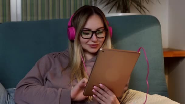 Una ragazza sul divano giace con un cane nel suo computer portatile e ascolta la musica attraverso le cuffie. Freelance e comunicazione via Internet — Video Stock