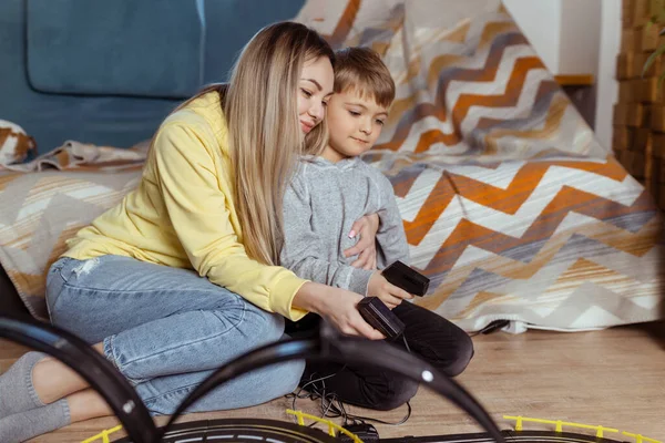 Mom and little son play racing on the carpet at home, have fun and hug. Single mother raises her son by playing cars