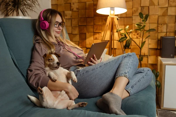 Aantrekkelijk meisje luistert naar muziek en kijkt videoclips op een tablet als een hond in een thuisomgeving binnen — Stockfoto