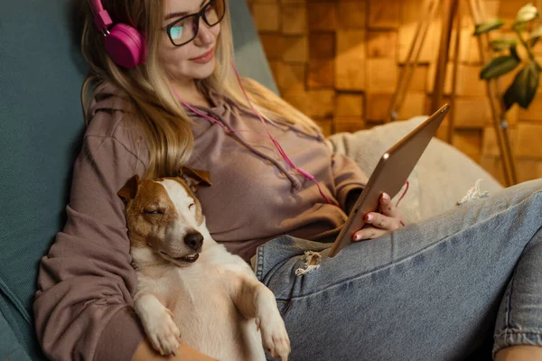 Een meisje op de bank ligt met een hond in haar laptop en luistert naar muziek via een koptelefoon. Freelancen en communicatie via internet — Stockfoto
