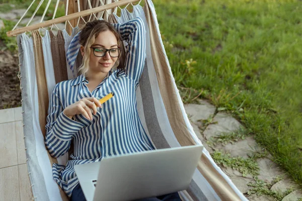 ハンモックの中の若い大人の女の子は家の外のコンピュータで働き電子タバコを吸う — ストック写真