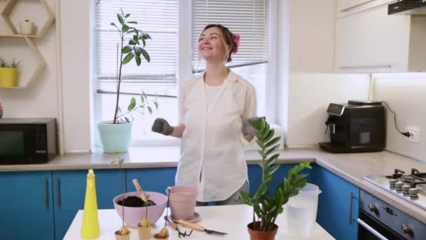 Uma menina bonito transplantes uma planta em casa enquanto dança engraçado em luvas de jardinagem. Caminhe e decore sua casa com plantas e flores frescas — Vídeo de Stock