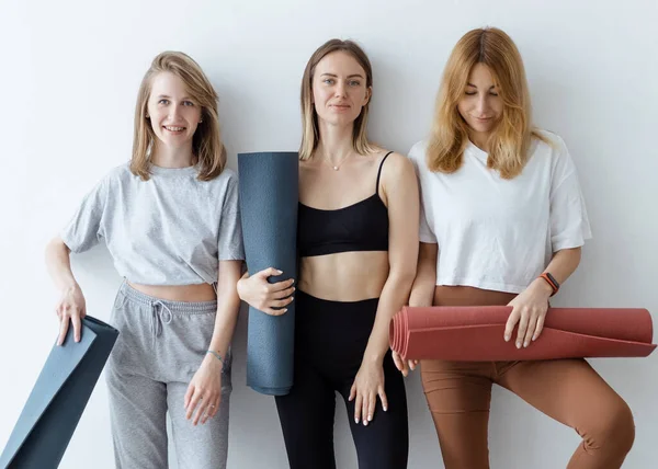 A group of young sports girls with yoga mats standing against a white wall. Girlfriends in the gym relaxing after fitness or yoga, indoors.