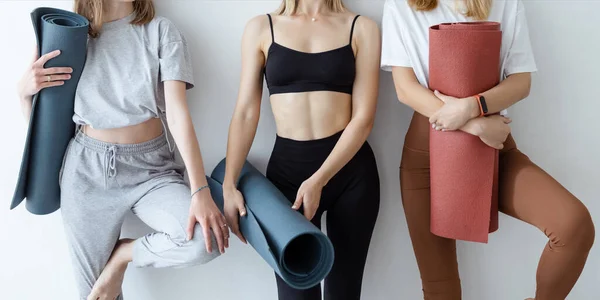 A group of young sports girls with yoga mats standing against a white wall. Girlfriends in the gym relaxing after fitness or yoga, indoors.