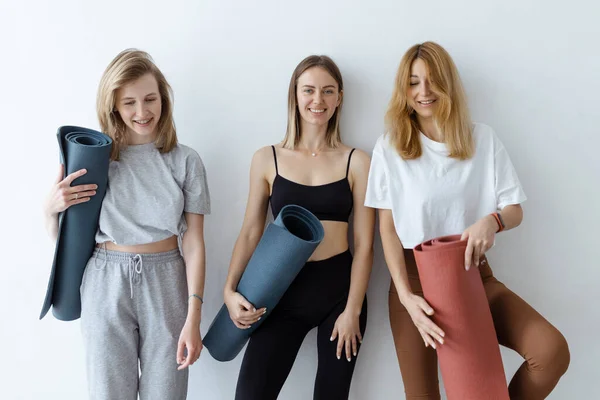 A group of young sports girls with yoga mats standing against a white wall. Girlfriends in the gym relaxing after fitness or yoga, indoors.
