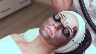 Güzellik uzmanları, güzellik salonundaki bir kanepede uzanan gözlüklü güzel bir müşteriye karbon soyuyorlar. Yüz temizleme konsepti. Kozmetik Hizmetleri. 