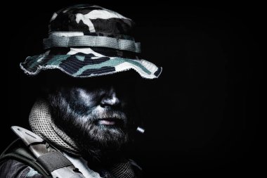 Yakın çekim, sakallı komando savaşçı düşük anahtar portre, ordu özel kuvvetler asker, boonie şapka özel askeri şirket paralı asker, taktik radyo kulaklık, siyah ve yeşil yüz kamuflaj boya