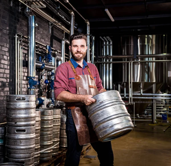 Jonge mannelijke brouwer in lederen schort houdt vat met ambachtelijk bier in moderne brouwerij fabriek — Stockfoto