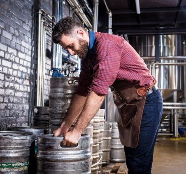 Deri önlüklü genç bira imalathanesinde bira fıçısı tutuyor.