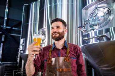 Deri önlük giyen genç bira imalathanesinde bira test ediyor.