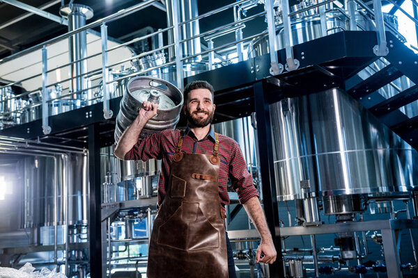 Молодой пивовар в кожаном фартуке держит бочку с ремесленным пивом на современном пивоварне