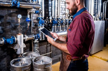 Modern bira fabrikasında bira fermantasyonunu denetleyen deri önlüklü genç bir erkek bira üreticisi.