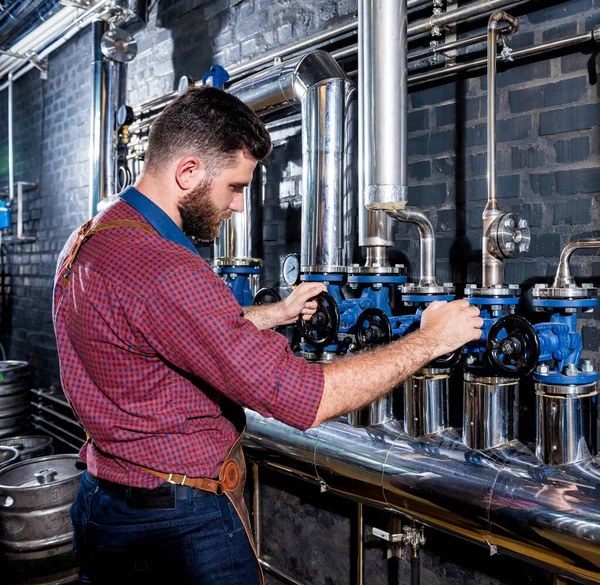 Jovem cervejeiro macho em avental de couro supervisionando o processo de fermentação da cerveja na fábrica moderna de cervejaria — Fotografia de Stock