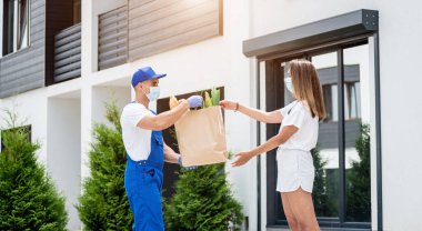 Koruyucu maske takan ve eldiven takan genç bir kurye karantina sırasında genç bir kadına mal teslim ediyor.