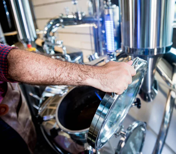 Fiatal, bőrkötényes férfi sörfőző, aki felügyeli a sörerjesztés folyamatát a modern sörgyárban — Stock Fotó