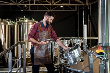 Modern bira fabrikasında bira fermantasyonunu denetleyen deri önlüklü genç bir erkek bira üreticisi.