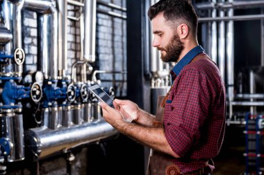 Modern bira fabrikasında bira fermantasyonunu denetleyen deri önlüklü genç bir erkek bira üreticisi.