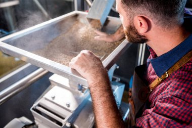 Deri önlüklü genç bir bira üreticisi modern bir bira fabrikasında malt tohumlarının öğütülmesini kontrol eder.
