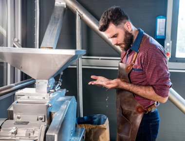 Deri önlüklü genç bir bira üreticisi modern bir bira fabrikasında malt tohumlarının öğütülmesini kontrol eder.