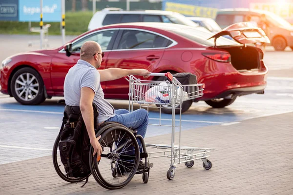 Person med en fysisk funktionsnedsättning skjuter en vagn mot en bil på en stormarknad parkeringsplats — Stockfoto