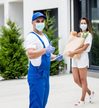 Koruyucu maske takan ve eldiven takan genç bir kurye karantina sırasında genç bir kadına mal teslim ediyor.