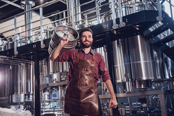 Молодой пивовар в кожаном фартуке держит бочку с ремесленным пивом на современном пивоварне