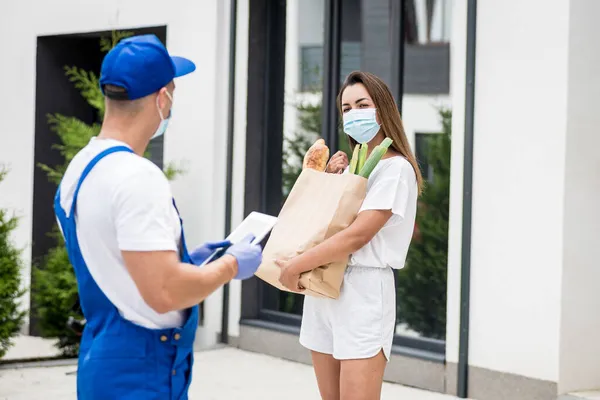 Un joven mensajero con una máscara protectora y guantes entrega mercancías a una joven durante la cuarentena. — Foto de Stock