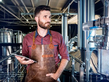 Modern bira fabrikasında bira fermantasyonunu denetleyen deri önlüklü genç bir erkek bira üreticisi.