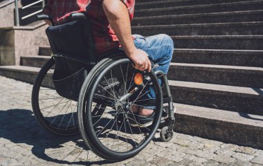 Merdivenlerin önünde tekerlekli sandalye kullanan fiziksel engelli genç bir adam.