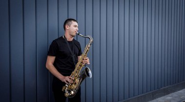 Genç sokak müzisyeni büyük mavi duvarın yanında saksafon çalıyor.