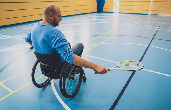 Дорослий чоловік з фізичною вадою, який використовує інвалідне крісло, граючи в теніс на критому тенісному корті — стокове фото
