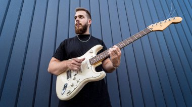 Büyük mavi duvarın yanında gitar çalan genç bir sokak müzisyeni.