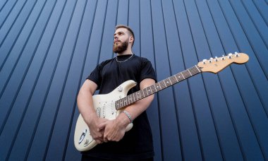 Büyük mavi duvarın yanında gitar çalan genç bir sokak müzisyeni.