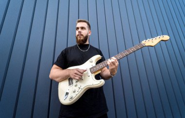 Büyük mavi duvarın yanında gitar çalan genç bir sokak müzisyeni.