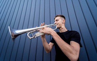 Büyük mavi duvarın yanında trompet çalan genç bir sokak müzisyeni.